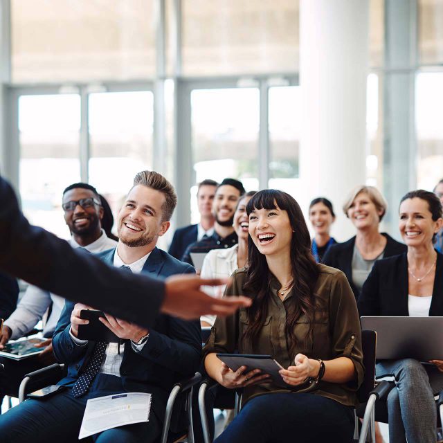 Professionals sitting in a conference setting smiling and laughing at a presenter.