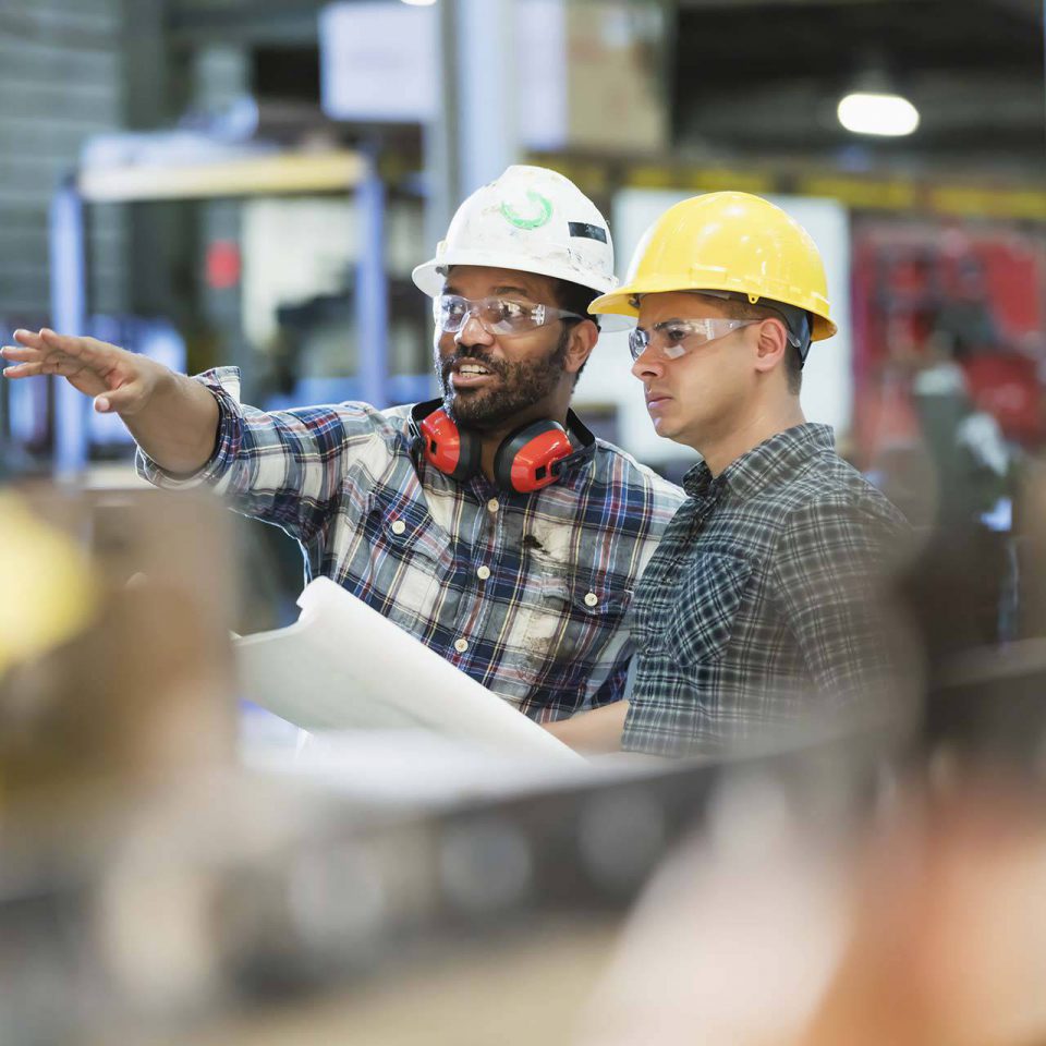 Two construction workers wearing hard hats talking while looking at blueprints