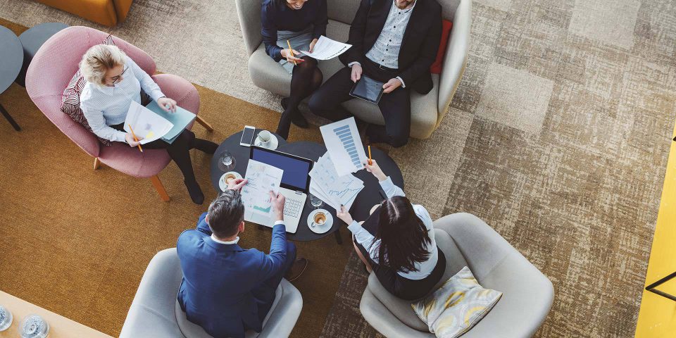 Aerial view of five professional adults sitting and networking over a laptop and paper handouts.